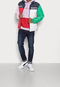 Padded jacket w kolorowych blokach w zielonym, czerwonym, białym i granatowym; czarny napis na klatce piersiowej. Nałożona na różową bluzę z kapturem, zestawiona z ciemnymi dżinsami i białymi sneakersami.