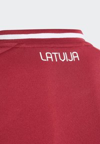Maillot de sport rouge avec des accents blancs, comportant "LATVIJA" en lettres majuscules au dos, fabriqué à partir d'un tissu léger et lisse.