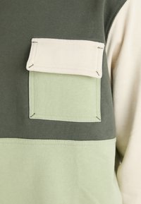 Camiseta de manga larga con un diseño en bloques de color en verde oscuro, verde claro y crema, con un bolsillo de parche y costuras contrastantes.