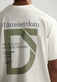 Camiseta de algodón color crema con texto gráfico en verde y azul oscuro que incluye detalles relacionados con los viajes y un diseño de "D" sobredimensionado en la parte posterior.
