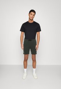 Zwarte T-shirt met korte mouwen, gecombineerd met donkergroene shorts met trekkoorden. Model draagt witte sneakers en enkelsokken, staand tegen een neutrale achtergrond.