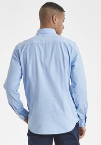 Camisa de manga larga de color azul claro en tejido ligero con cuello abotonado, dobladillo curvado y acentos de botones en contraste en los puños.
