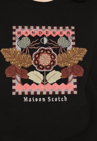 Prenda negra con bordado colorido, que representa hojas, flores y el texto "WANDERER" y "Maison Scotch" en diferentes texturas.