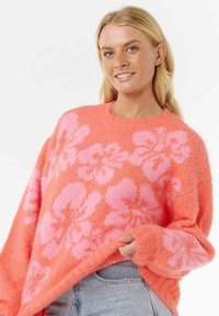 Frau mit blonden Haaren lächelt, trägt einen lockeren, flauschigen korallenfarbenen Pullover mit großen pinken Blumenmustern und hellblauen Jeans.