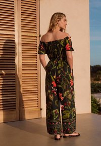 Salopetă florală cu umeri descoperiți, din material negru, cu frunze verzi și flori vibrante, având talie elastica și picioare largi.