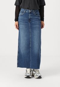 Jupe en jean maxi bleue foncée, avec une coupe droite, une fente avant et un ourlet brut. Associée à des baskets argentées et un haut noir.