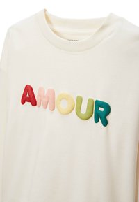 T-shirt en coton couleur crème avec le mot "AMOUR" en lettres colorées, texturées et en relief. Les lettres varient en couleur : rouge, rose, jaune, vert, bleu.