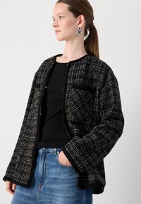 Mustvalge tweed jakk, millel on kasti kujuline lõige, avatud esikülg, kaks esikotid, laiad varrukad ja tekstuurilised detailid. Kätkse musta särgi peal.