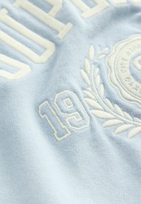 Ljust blått tyg med vit broderi som visar texten "SUPER" och siffran "19," prydd med en lagerkransdesign.