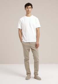 Camiseta blanca de algodón, de manga corta, con cuello redondo, combinada con pantalones de algodón beige claros y zapatillas slip-on beige, sobre un fondo neutral.