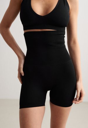 Kvinne iført høytlivede svarte formende shorts og en svart sports-BH, stående med en hånd på hoften mot en nøytral bakgrunn.