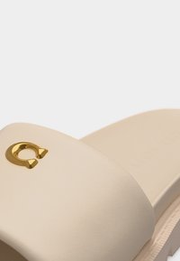 Μπεζ σανδάλι με φαρδιά λουρί που διαθέτει μικρό μεταλλικό διακοσμητικό σε σχήμα πετάλου από χρυσό στο επάνω μέρος.