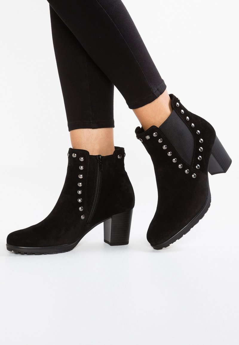 Czarne zamszowe botki do kostki z srebnymi ćwiekami, na słupku, z bocznym zamkiem błyskawicznym i elastycznymi panelami, noszone z czarnymi dopasowanymi spodniami.