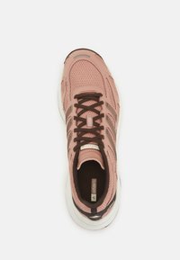 Baskets d'athlétisme rose clair avec tige en mesh, lacets marron et accents foncés. Présentent une semelle blanche et des détails métalliques sur le côté.