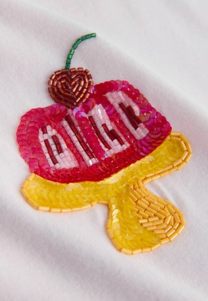 Spīguļi izšuvums ar saldējuma konusa dizainu, sarkanu saldējuma bumbiņu, dzeltenu konusa daļu un zaļu ķirsi uz balta auduma fona.