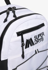 Sac à dos en nylon blanc avec des accents noirs, présentant des doubles zips, un marquage et des cordons élastiques. Texture lisse et design structuré.