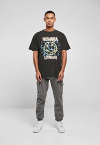 Juodas per didelis T-shirt su "NIRVANA" grafika, daugiaspalviais akcentais. Suderinta su šviesiai pilkomis kargo kelnėmis ir baltomis sportinėmis bateliais.