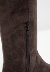 Braune Wildlederstiefel bis zum Knie mit einem schwarzen Reißverschlussdetail an der Seite. Glatte Textur mit passgenauer Silhouette.