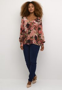 Blomstret bluse med lange ærmer i pink og grønne nuancer, parret med mørke skinny jeans og beige højhælede sko, sat mod en neutral baggrund.