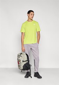 Λαχανί αθλητική μπλούζα με κοντά μανίκια, γκρι φόρμα με πλαϊνές λεπτομέρειες και ανοιχτογκρι σακίδιο με διχτυωτό σχέδιο. Μαύρα αθλητικά παπούτσια.