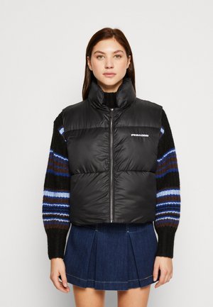 Schwarze Steppweste mit hohem Kragen, versehen mit einem Reißverschluss vorne und einem Logodetail. Kombiniert mit einem gestreiften Strickpullover und einem Jeansrock.