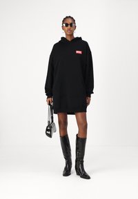 Vestido hoodie preto com um bolso frontal e o logo 'DIESEL' em vermelho. Usado com botas pretas brilhantes até ao joelho e segurando uma pequena mala preta.