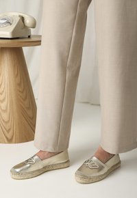 Espadrilles metálicas doradas con suela de yute trenzada, que presentan un diseño bordado en la puntera. Acompañadas de pantalones beiges ajustados.