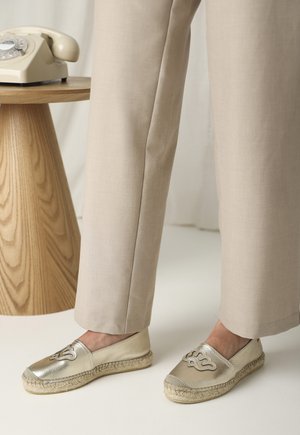 Espadrillas dorate metallizzate con suola in juta intrecciata, caratterizzate da un design ricamato sulla punta. Abbinato a pantaloni beige su misura.