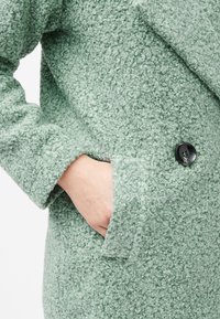 Cappotto verde texturizzato realizzato in un tessuto morbido e peloso, con colletto a scialle e tasca laterale con dettaglio in bottone nero.
