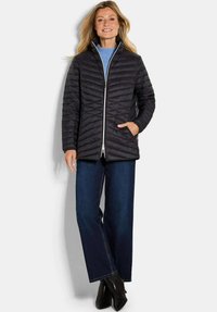 Woman wearing a black quilted jacket, blue sweater, dark jeans, and black ankle boots, standing with one hand in pocket and smiling.