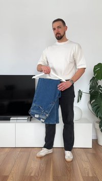 Biała koszulka z krótkim rękawem noszona przez osobę trzymającą niebieskie dżinsy na białym wieszaku, z ciemnymi spodniami i beżowymi sneakerami, w jasnym pomieszczeniu.