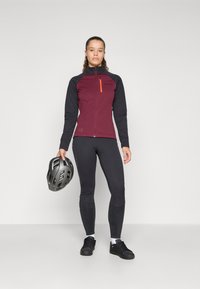 Bordeaux en zwarte fietsjack met hoge kraag, ritssluiting aan de voorkant, oranje accenten en zwarte leggings. Model houdt een grijze helm vast.