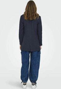 Sacou bleumarin dintr-un material texturat, cu mâneci lungi și slit la spate, asortat cu pantaloni largi din denim albastru și adidași albi cu accente negre.