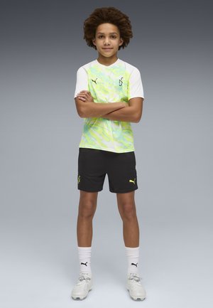 Niño de pie con los brazos cruzados, vestido con una camiseta deportiva con patrón neón, pantalones cortos negros, calcetines blancos y zapatillas blancas sobre un fondo gris.