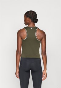 Γυναίκα με πλεγμένα μαλλιά, φορώντας ελιά πράσινη αμάνικη μπλούζα και μαύρα κολάν, που στέκεται με πλάτη σε έναν απλό φόντο.