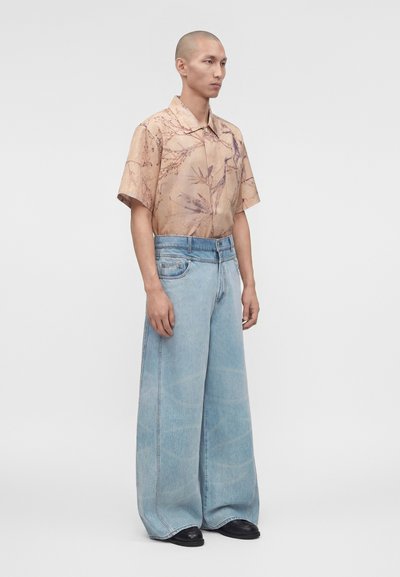 Άνδρας που φοράει μια μπεζ κοντομάνικη πουκάμισο με σχέδιο, κρυμμένη μέσα σε ψηλόμεσο, ανοιχτό μπλε παντελόνι με φαρδύ πόδι και μαύρα παπούτσια.