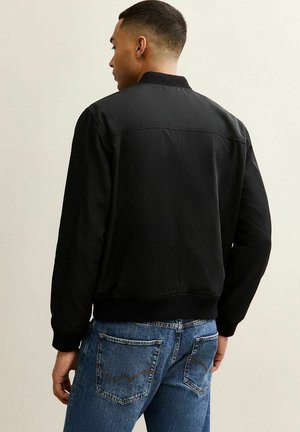 Młody mężczyzna z krótkimi włosami, ubrany w czarną kurtkę bomberkę i niebieskie dżinsy, stoi bokiem na jasnym, jednolitym tle.