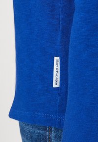 Sininen puuvillapaita, jossa on sileä rakenne, ja sivussa on etiketti, jossa lukee "Marc O'Polo DENIM." Helma on siististi ommeltu.