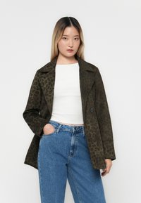Grön leopardmönstrad kappa med notchad krage, enkel knappstängning och sidofickor, kombinerad med en vit topp och blå jeans.