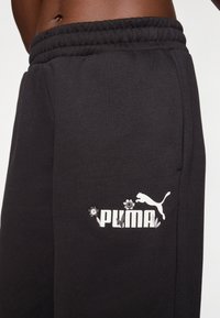 Black joggers with an elastic waistband, featuring a white Puma logo and floral accents printed near the left pocket. Soft texture.