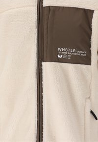 Chaqueta de forro polar en color crema con detalles en marrón, con cierre de cremallera y bolsillo lateral. Incluye una marca que dice "WHSTLR" y "Ultimate Protective Wear".