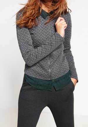 Kvinde iført en tekstureret grå lynlåstrøje over en mørkegrøn skjorte med krave og mørke bukser, med den ene hånd i lommen og den anden nær kraven.