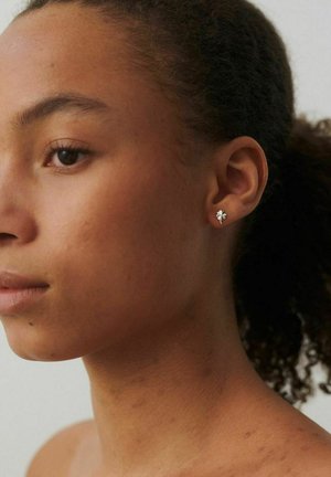 Lähikuva kiharatukkaisesta henkilöstä, jolla on pienet hopeiset nelilehtiset apilan muotoiset korvakorut vasemmassa korvassa.