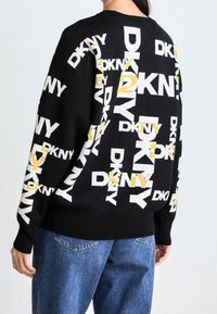 Svart stickad tröja med ett upprepat logomönster i vitt och gult. Den har en avslappnad passform, sänkta axlar och ribbade muddar.