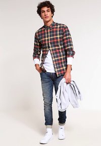 Geruit flanellen overhemd in marineblauw, rood en crème, met een wit ondershirt. Gecombineerd met een blauwe spijkerbroek en witte sneakers, terwijl een grijze sweatstof vastgehouden wordt.