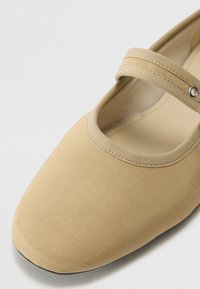 Zapato tipo Mary Jane en lona beige con punta redonda, tira elástica y superficie texturizada. Incluye un acento metálico circular cerca de la tira.