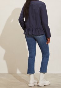 Námořnický peplum top s dlouhými rukávy, spárovaný s modrými skinny džíny a bílými masivními kotníkovými botami, zobrazený zezadu na neutrálním pozadí.