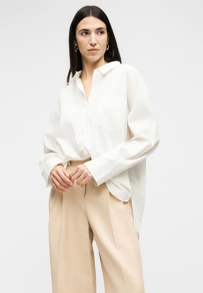 Femme aux longs cheveux foncés portant des boucles d’oreilles créoles dorées, une chemise blanche oversize boutonnée et un pantalon beige à jambes larges sur un fond uni.