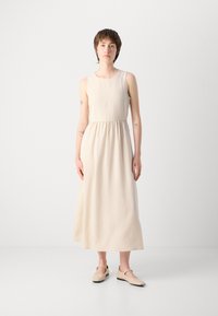 Light beige sleeveless dress with a gathered waist, reaching mid-calf, made of smooth fabric; paired with beige slip-on shoes.