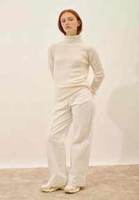Kremowy sweter z prążkowanym kołnierzem typu golf, zestawiony z szerokimi białymi spodniami i beżowymi sneakerami. Gładka tekstura i luźny krój, z subtelnymi detalami w stichach.
